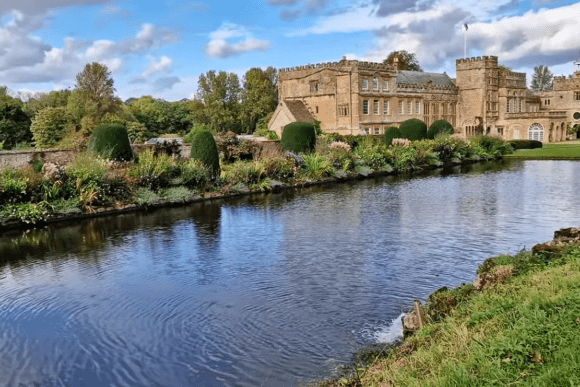 Forde Abbey reflected in a tranquil pond, with manicured gardens and historic stone buildings under a partly cloudy sky.