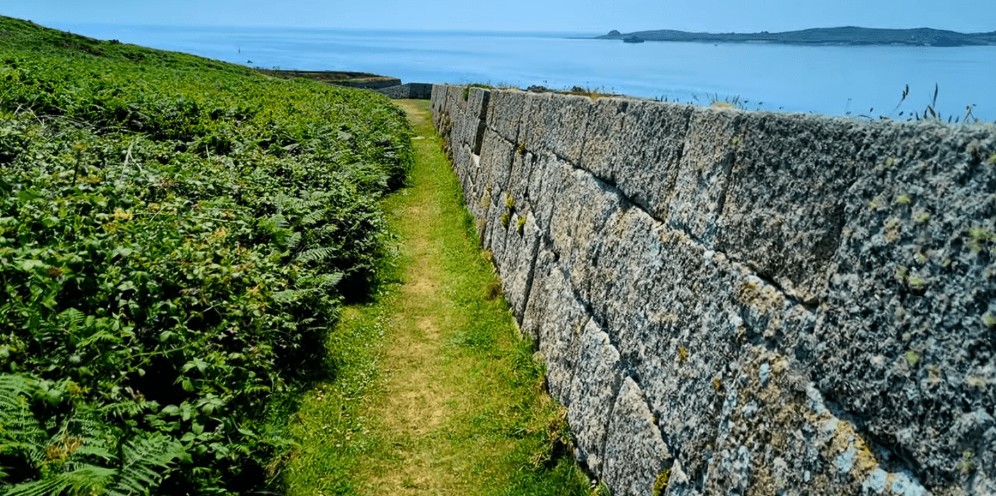 Narrow grassy path running alongside a stone garrison wall with dense greenery on one side and the sea visible beyond.