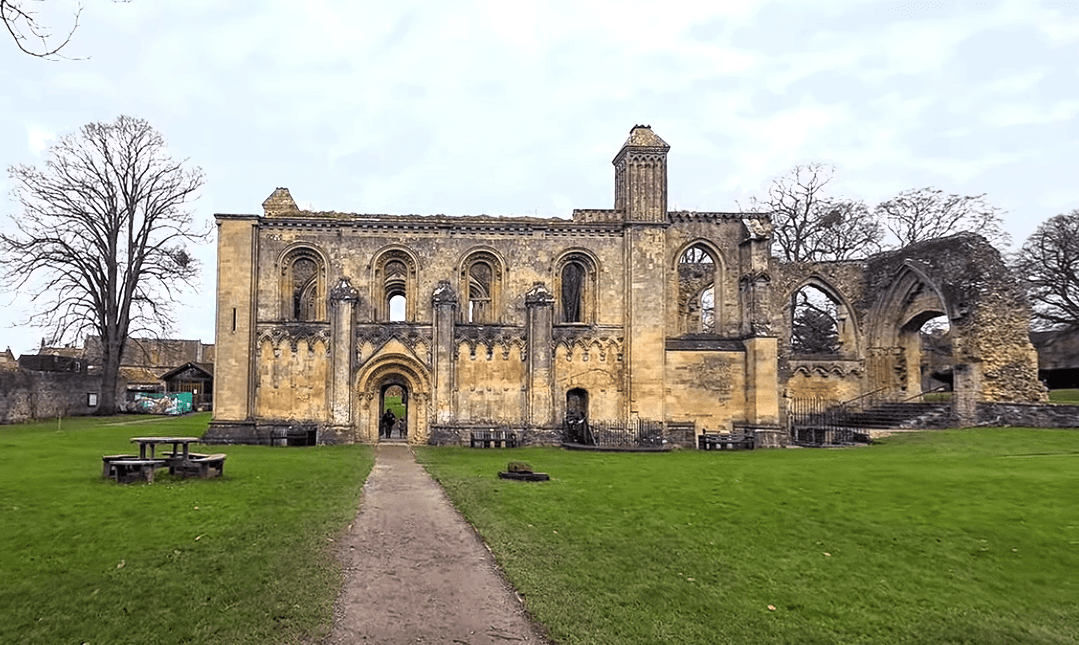 Front view of the surviving west front of Glastonbury Abbey ruins with a central arched doorway, tall empty window openings, and a grassy lawn in the foreground.