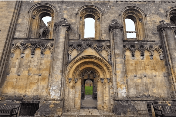 Close-up view of Glastonbury Abbey’s west front showing an ornate Romanesque arched doorway, decorative stone carvings, tall empty window openings, and weathered medieval masonry.