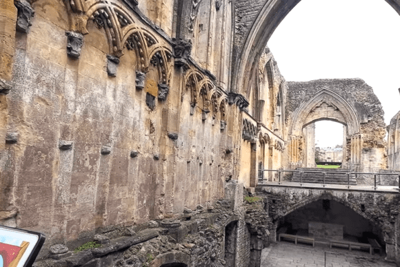 Interior view of Glastonbury Abbey ruins showing tall Gothic arches, carved stone arcading, and a stone undercroft beneath a raised walkway.