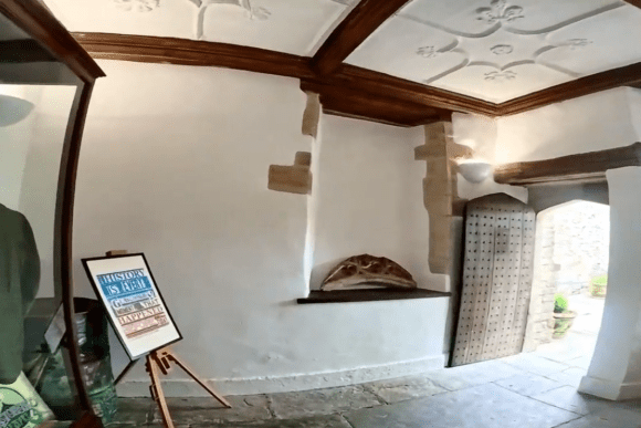 Interior of the Glastonbury Tribunal showing white plaster walls, timber ceiling beams, a display case, and an open studded wooden door leading outside.
