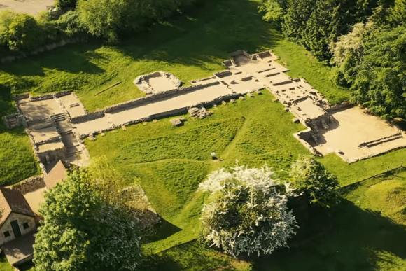 Aerial view of the ruins of Great Witcombe Roman Villa showing stone foundations and room outlines set within green countryside.