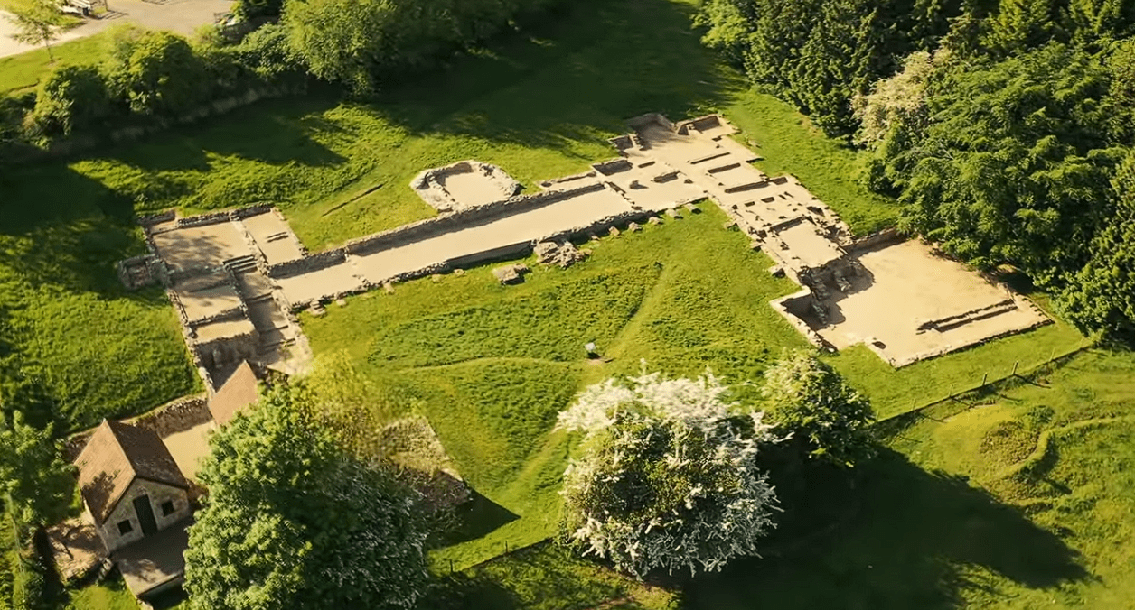 Aerial view of the ruins of Great Witcombe Roman Villa showing stone foundations and room outlines set within green countryside.