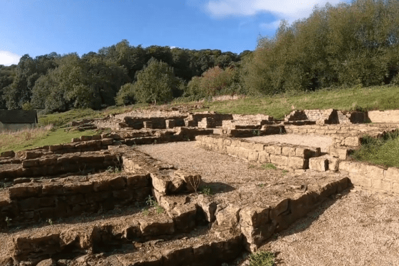 Stone foundations and low walls of Great Witcombe Roman Villa ruins set on a grassy hillside with trees in the background.