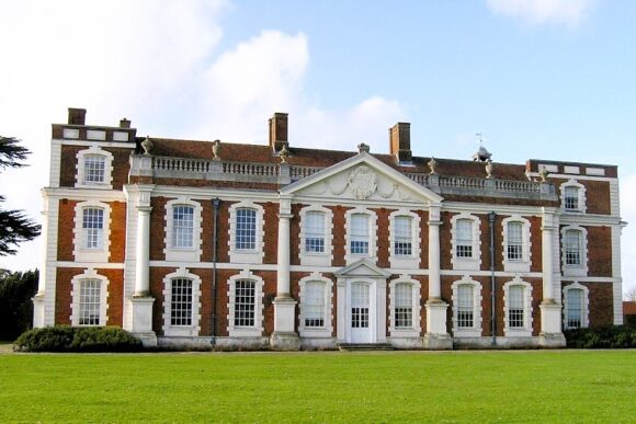 Front view of Hill Hall, a historic red-brick Elizabethan mansion with white stone columns and large windows.