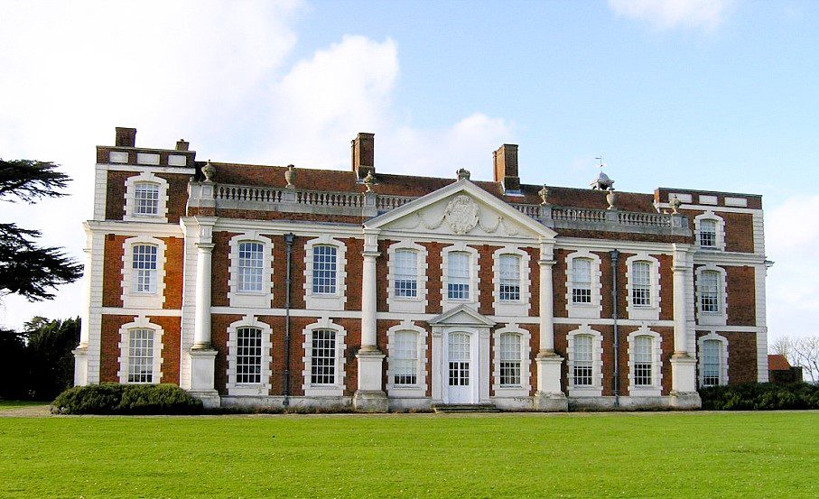 Front view of Hill Hall, a historic red-brick Elizabethan mansion with white stone columns and large windows.