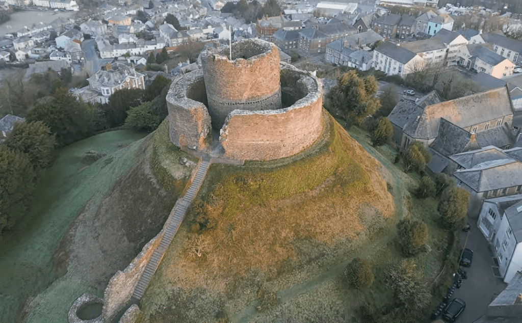 Aerial view of Launceston Castle perched on a grassy mound, showing its circular stone walls and central keep surrounded by a town.