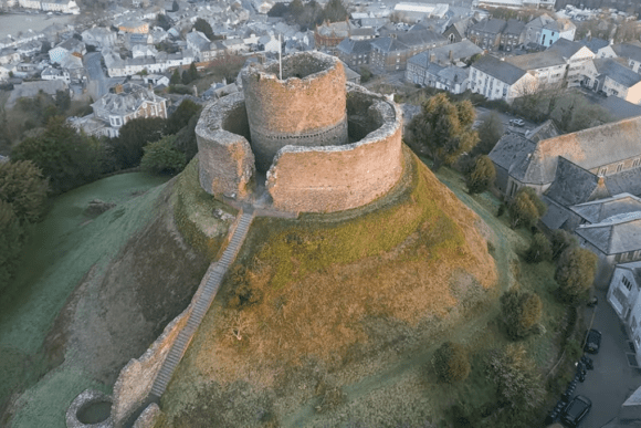 Aerial view of Launceston Castle perched on a grassy mound, showing its circular stone walls and central keep surrounded by a town.