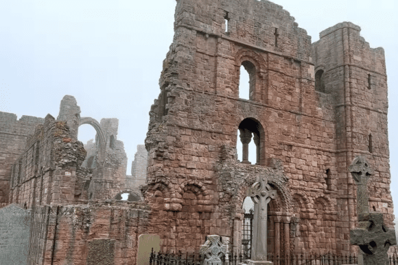 Weathered red stone ruins of Lindisfarne Priory with arched openings and Celtic gravestones in the foreground.