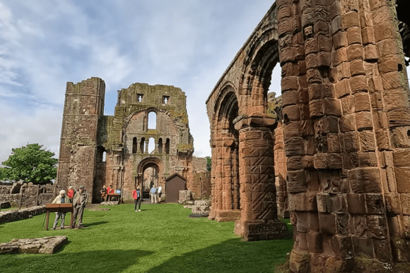 Ruins of Lindisfarne Priory with arched stone columns, visitors on grass, and a large historic facade in the background.