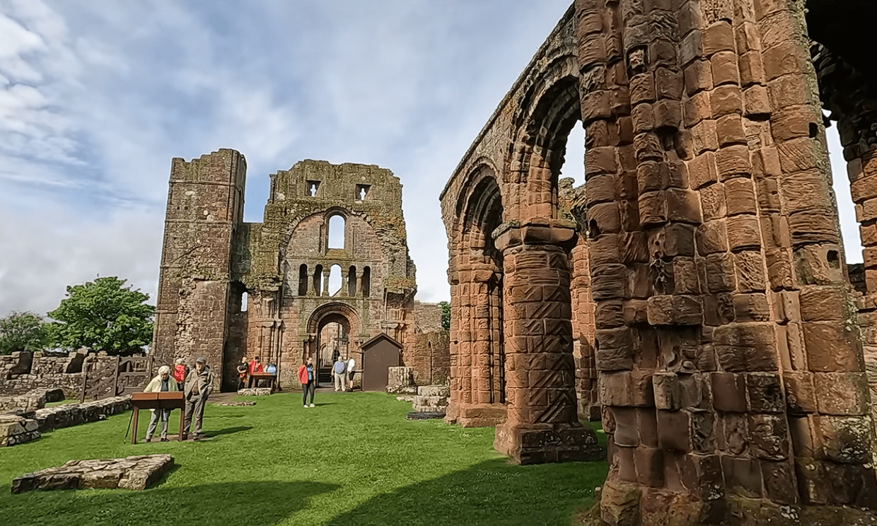 Ruins of Lindisfarne Priory with arched stone columns, visitors on grass, and a large historic facade in the background.