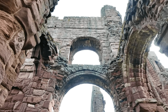 View looking up at red sandstone arches and walls inside Lindisfarne Priory ruins against the sky.