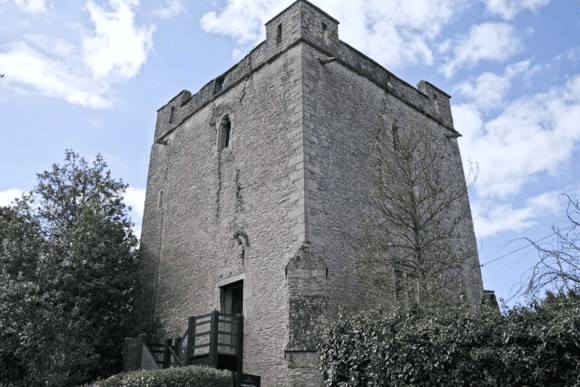 Stone tower of Longthorpe Tower surrounded by trees and greenery under a partly cloudy sky.