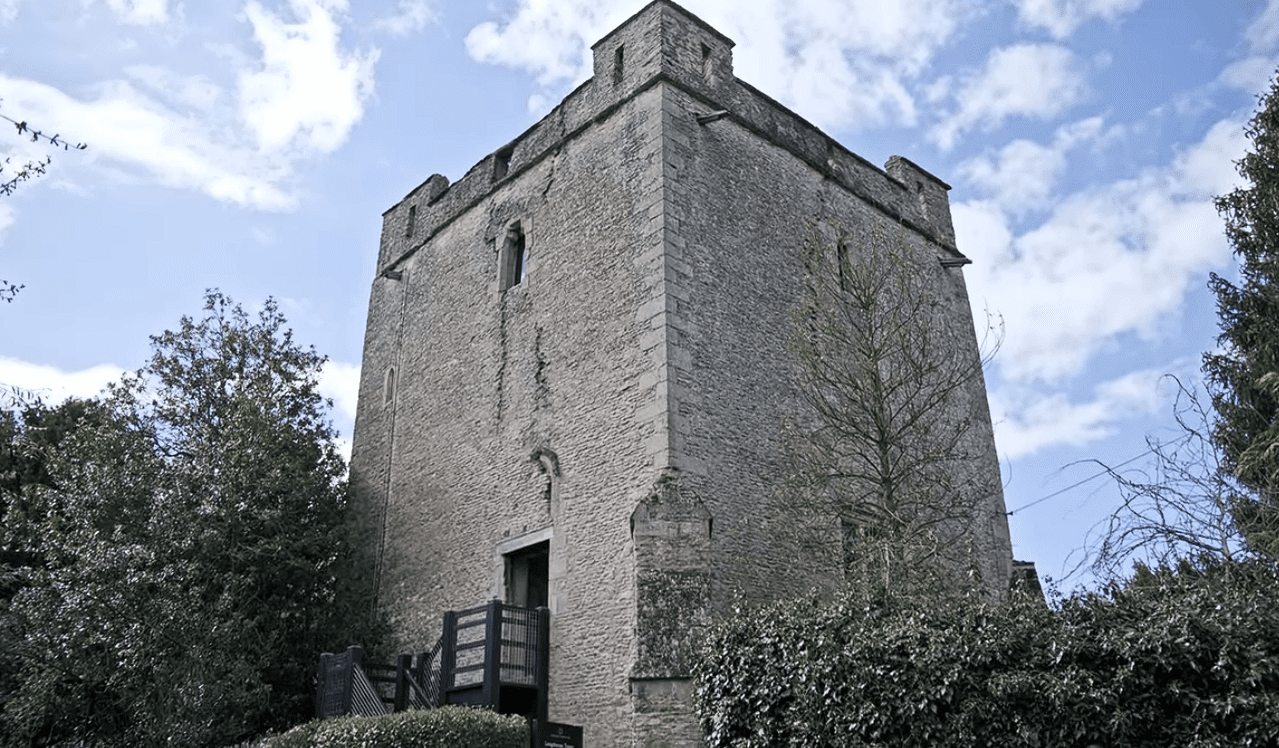 Stone tower of Longthorpe Tower surrounded by trees and greenery under a partly cloudy sky.