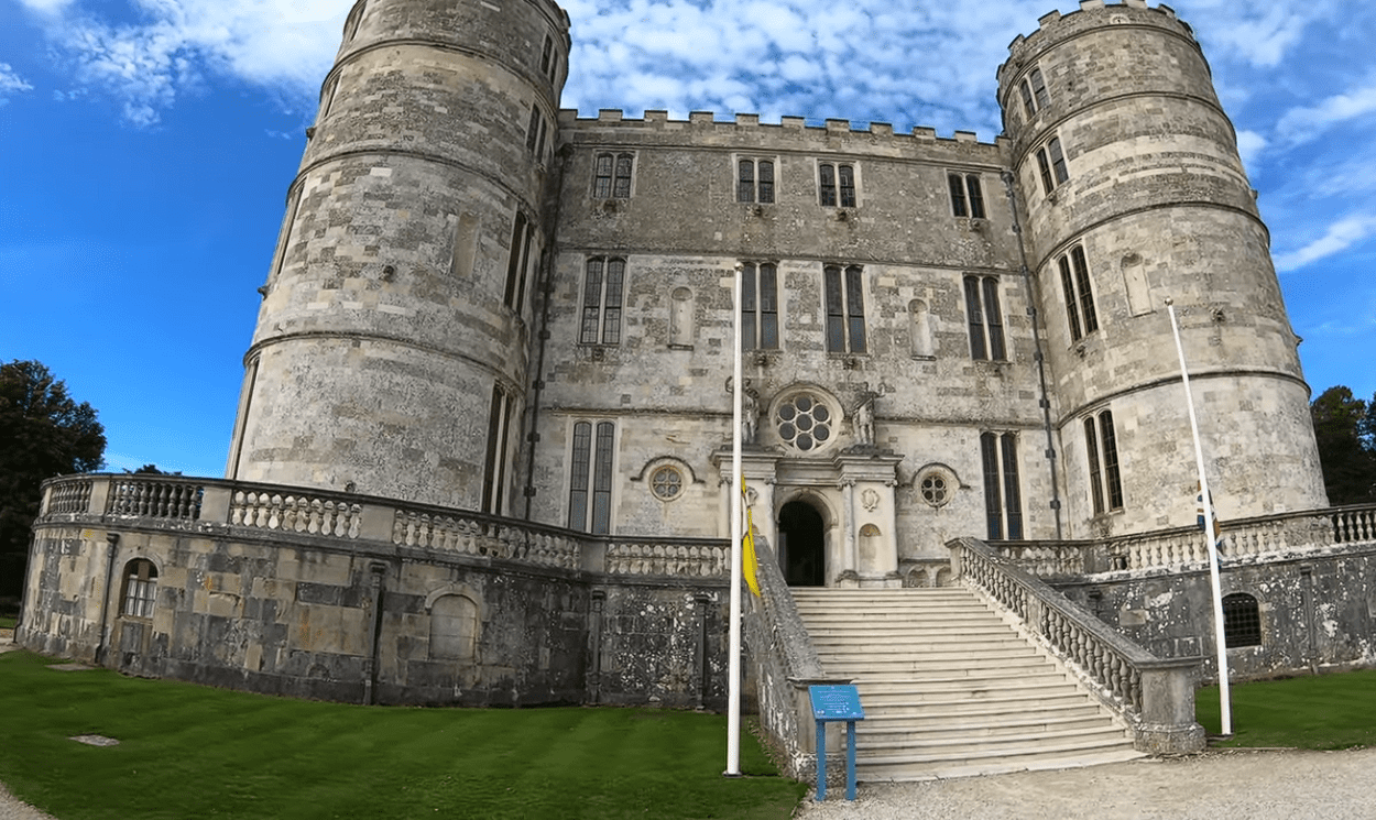 Front view of Lulworth Castle with twin round towers, stone steps, and entrance under a blue sky.