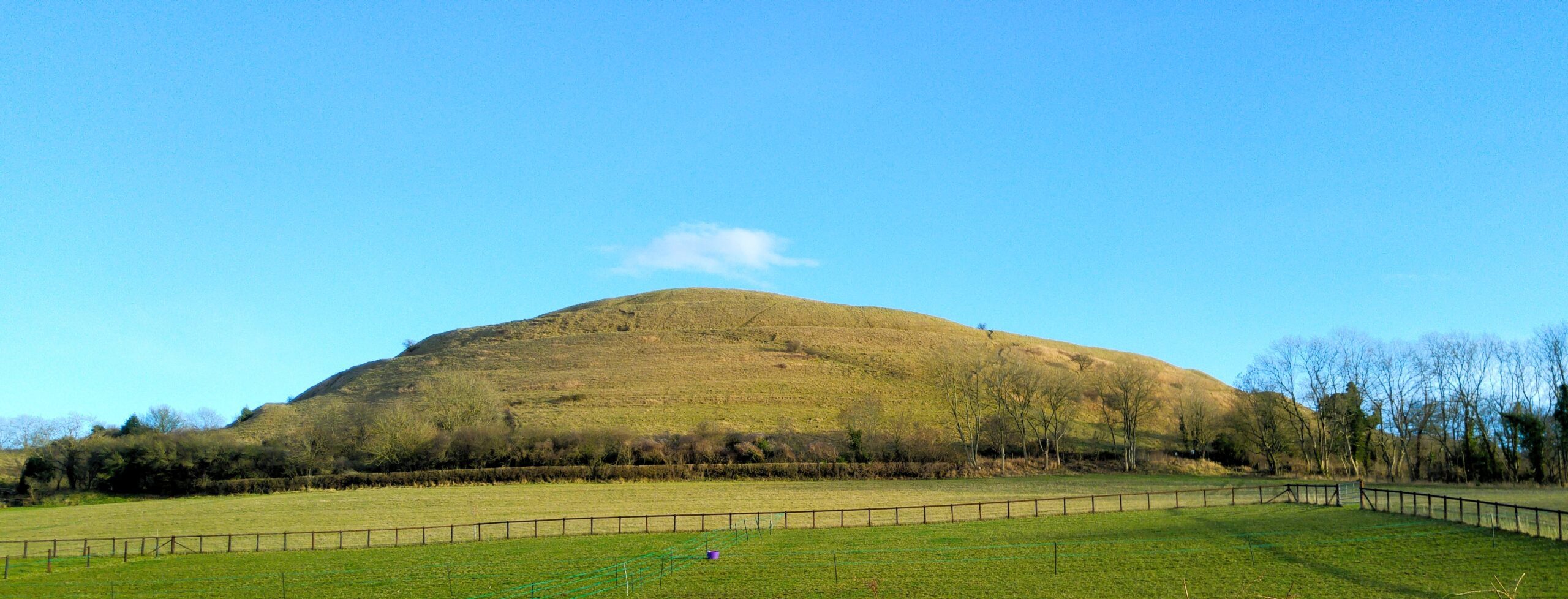 Hambledon Hill rising above green fields under a clear blue sky in Dorset.