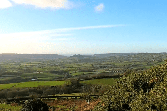 Panoramic view from Pilsdon Pen in Dorset, England, overlooking rolling green countryside, patchwork fields, and distant hills under clear blue skies.