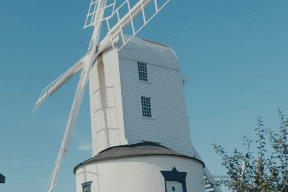 Saxtead Green Post Mill in Suffolk, traditional white wooden windmill with sails against a clear blue sky.
