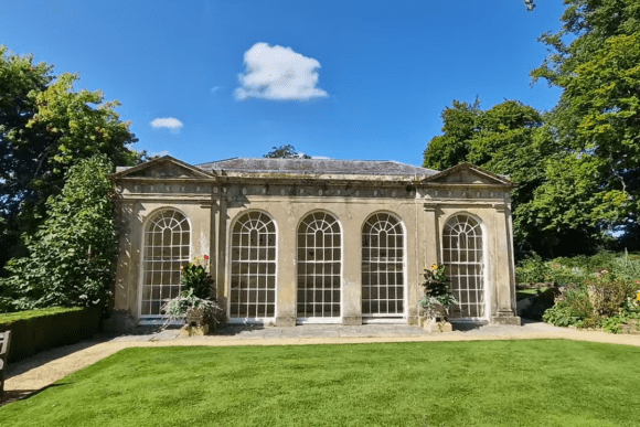 Orangery building at Sherborne Old Castle gardens with arched windows, manicured lawn, and surrounding greenery under blue sky.