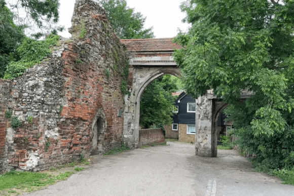 Historic Waltham Abbey Gatehouse and medieval stone bridge surrounded by trees, featuring Gothic arches, weathered brick walls, and a quiet pathway in Essex, England.