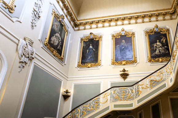 Grand staircase hall inside Wrest Park featuring ornate gold-framed portraits, decorative wrought iron railings, and elegant Georgian interior design.