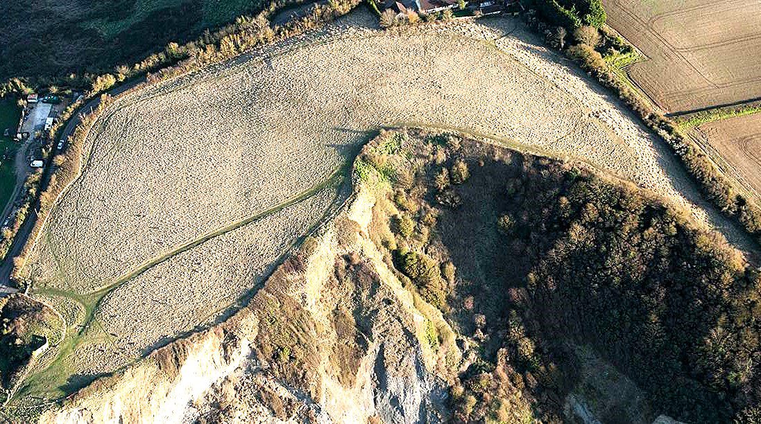 Aerial view of Daw’s Castle earthworks on a clifftop, showing curved defensive banks above steep coastal slopes.