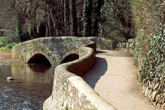 Curved stone packhorse bridge with twin arches over a shallow river at Dunster Gallox Bridge.