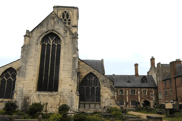 Ruins of Greyfriars church with tall Gothic windows beside historic red-brick buildings and a small churchyard.