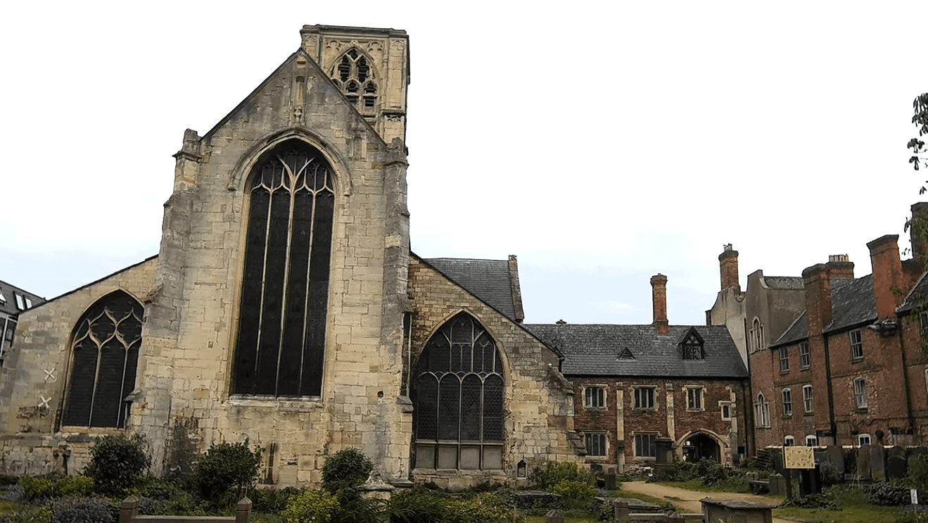 Ruins of Greyfriars church with tall Gothic windows beside historic red-brick buildings and a small churchyard.