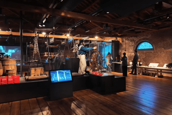 Interactive maritime history gallery inside the Museum of London Docklands featuring historic shipping equipment, cargo-handling tools, a sailor figure at a ship’s wheel, and immersive docklands exhibits within a restored warehouse setting.