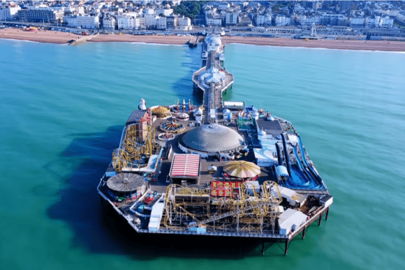 Aerial view of Brighton Palace Pier extending into the sea, with amusement rides, Brighton Beach and the city seafront in the background.
