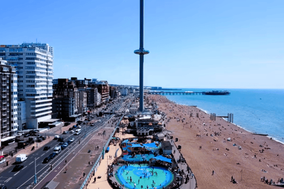 Panoramic view of Brighton Beach with the Brighton i360, seafront promenade, beachgoers and Brighton Palace Pier beside the sea.