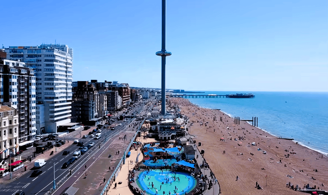 Panoramic view of Brighton Beach with the Brighton i360, seafront promenade, beachgoers and Brighton Palace Pier beside the sea.
