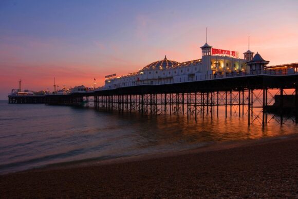 Brighton Palace Pier illuminated at sunset, extending over the sea from Brighton Beach beneath a colourful pink and orange sky.